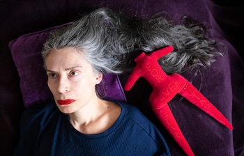 Retrato de Penny Goring recostada sobre una superficie púrpura, con cabello gris y labios rojos, junto a una figura de tela roja con la inscripción 'FOREVER LIAR'