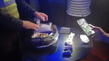 Agente policial inspecciona dinero y monedas en una mochila, junto a paquetes rectangulares blancos y azules sobre una mesa oscura. Una persona sostiene un teléfono móvil
