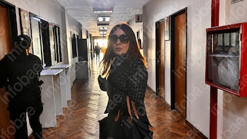 Mujer con gafas de sol y abrigo oscuro en un pasillo largo de piso de madera. Al fondo entra luz. Persona de seguridad de espaldas a la izquierda. Extintor en la pared