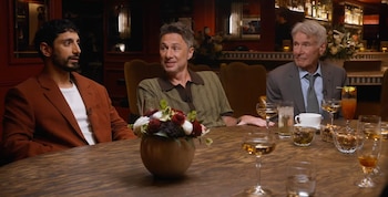 Tres hombres sentados en una mesa de madera. Harrison Ford, de traje gris, conversa con dos hombres. En la mesa hay bebidas y un adorno floral