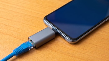 Un teléfono móvil conectado a un cable azul mediante un adaptador gris sobre una mesa de madera clara.