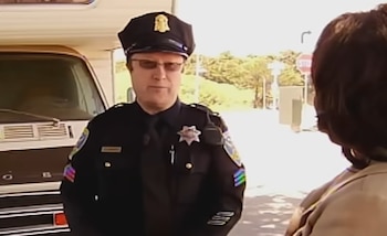 Un oficial de policía con gorra, gafas de sol y uniforme oscuro habla con una persona frente a un vehículo blanco