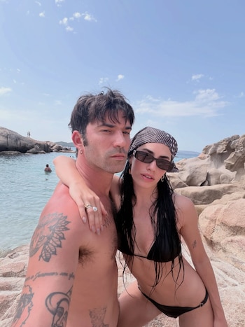 Un hombre sin camiseta y una mujer con bikini negro, gafas de sol y pañuelo en la cabeza posan en una playa rocosa con mar y cielo azul