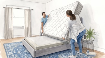 Un hombre y una mujer levantan un colchón blanco para ponerlo en una base de cama. Hay una ventana con cortinas, una alfombra, una planta, libros y un vaso.