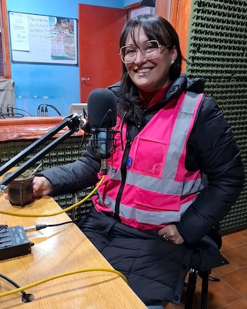 Una mujer con gafas y chaleco rosa de alta visibilidad sostiene un mate en una cabina de radio frente a un micrófono