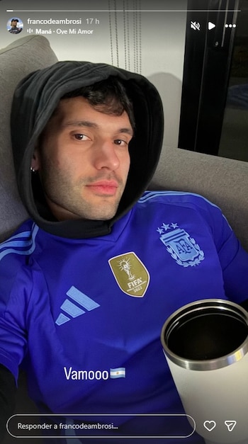 Hombre joven con hoodie negro y camiseta azul claro de la selección argentina con escudo de campeón FIFA 2022. Sostiene una taza blanca