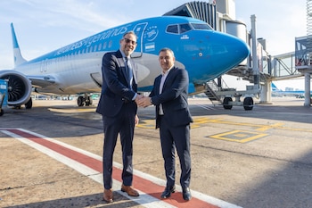 Dos hombres de traje y lentes se dan la mano delante de un avión azul y blanco de Aerolíneas Argentinas y una pasarela de embarque en una pista de aeropuerto