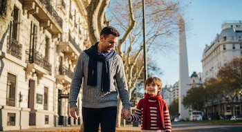 Padre e hijo, abrigados con bufanda y pulóver, caminan tomados de la mano por una calle de Buenos Aires bajo el sol. El Obelisco es visible al fondo.