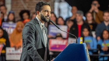 Zohran Mamdani, un hombre barbudo con traje gris y corbata, sonríe mientras habla en un micrófono en un podio azul, con una multitud borrosa detrás