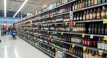 Vista de un largo pasillo de supermercado lleno de estanterías con cientos de botellas de vino y licores, con algunos clientes empujando carritos de compra al fondo.