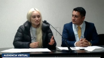 Mujer rubia con buzo plomo y casaca negra en audiencia virtual, junto a un abogado con traje azul, sentados ante una mesa con micrófono y papeles.