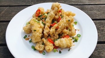 Plato blanco con calamares fritos a la sal y pimienta, de forma irregular y sin tentáculos. Guarnición de ajo frito, chile rojo y cebolletas, sobre mesa de madera oscura.