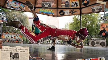 Ilustración tipo collage de un breakdancer en una pose dinámica, con ropa deportiva roja, en un pabellón. Alrededor hay grafitis, boomboxes y discos de vinilo.