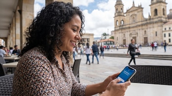 Una mujer con cabello rizado oscuro mira y sonríe a su teléfono en una terraza. Al fondo, se ve una gran catedral de piedra clara bajo un cielo nublado.