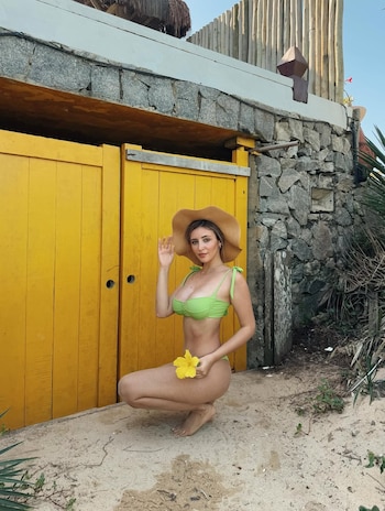 Abril Duhalde, en bikini verde y sombrero de paja, se agacha sobre arena. Sostiene una flor amarilla frente a puertas amarillas y una pared de piedra