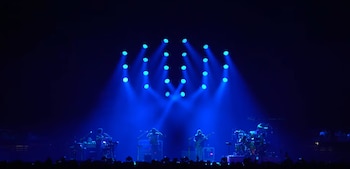 Cuatro músicos tocan instrumentos en un escenario iluminado con luces azules. Una audiencia se observa en la parte inferior