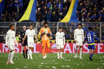 Portero de uniforme naranja y guantes negros en césped. Otros futbolistas con camisetas blancas y uno con camiseta azul. Árbitro. Público y banderas al fondo. Confeti en el suelo