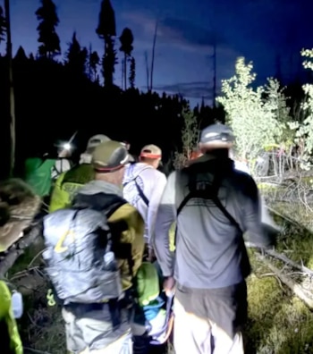 Grupo de personas con mochilas y linternas frontales camina por un sendero boscoso oscuro con árboles y vegetación