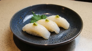 Tres piezas de Ika nigiri de calamar con una pequeña porción de wasabi verde en la parte superior, servidas en un plato oscuro texturizado, sobre una superficie clara y granulada.