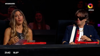 Una mujer de pelo rubio y un hombre con chaqueta militar azul, gafas de sol y cadena dorada sentados en una mesa oscura con luces rojas