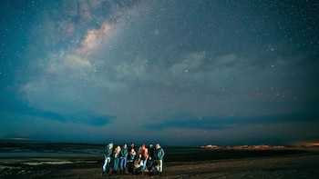 Un grupo de diez personas con ropa de abrigo posa de noche en la arena bajo un cielo estrellado con la Vía Láctea visible