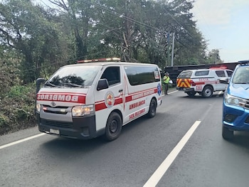 Ambulancia de Bomberos Voluntarios, una camioneta de rescate y un coche azul en una carretera; un vehículo grande, posiblemente un autobús, está volcado al fondo