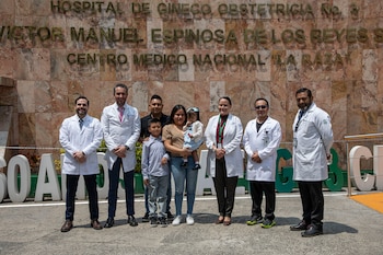 Un grupo de siete médicos de bata blanca posa junto a una familia de cuatro (dos adultos, dos niños) frente a la fachada de piedra de un hospital