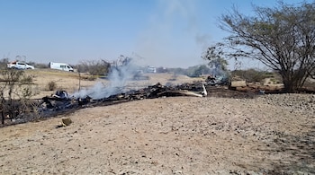 Escombros quemados de una aeronave sobre terreno árido con humo ascendente. A lo lejos se ven arbustos secos y vehículos de emergencia blancos