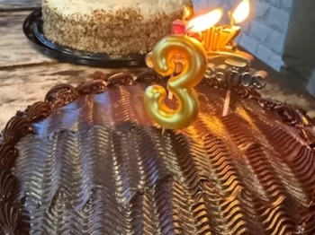 Pastel de chocolate con glaseado decorado, vela dorada en forma de número tres y otras velas encendidas. Un segundo pastel claro en segundo plano