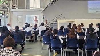Vista interior de un seminario con una mujer presentando a una audiencia sentada en mesas. Una pantalla proyecta texto en español y un póster con mensaje