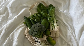 Vista aérea de una bolsa de malla beige llena de vegetales y frutas verdes frescos: brócoli, pepinos, aguacate, cilantro, bok choy y limas, sobre tela blanca.