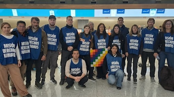 Un grupo de 16 personas, varias con chalecos azules que dicen 'Misión Derechos Humanos', posan en un interior moderno; una persona sostiene una bandera Wiphala.