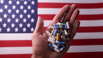 Close de uma mão segurando vários comprimidos e cápsulas de diferentes cores e tamanhos, com a bandeira dos Estados Unidos desfocada ao fundo.