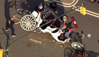 Vista aérea de un carruaje blanco y rojo volcado en una calle gris. Un hombre está de pie junto a una rueda, escombros dispersos y figuras cerca del vehículo