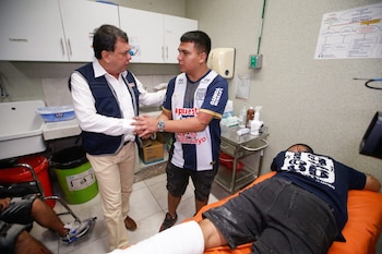 Hombre en chaleco azul habla con joven en camiseta de fútbol a rayas en una sala de hospital; otro paciente yace en camilla con pierna vendada