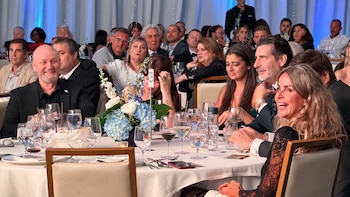 Un gran grupo de personas sentadas en mesas redondas en una gala, con copas de agua y vino, y centros de mesa florales. Luces azules de fondo