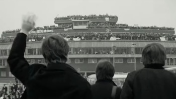 Imagen en blanco y negro de tres figuras masculinas vistas de espaldas, una de ellas saludando a una gran multitud de personas en las terrazas de un aeropuerto