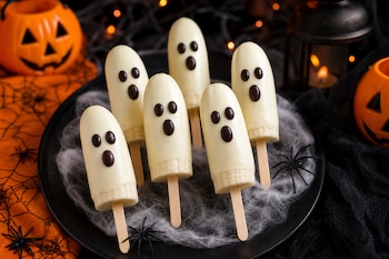 Seis trozos de plátano en palitos cubiertos de chocolate blanco con ojos de chocolate sobre un plato con telarañas y calabazas decorativas.