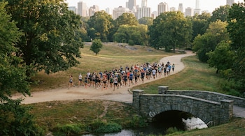 Vista aérea de un gran grupo de hombres y mujeres corriendo en un sendero de tierra en un parque, con un puente de piedra sobre un arroyo y edificios urbanos al fondo.