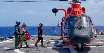 Un helicóptero rojo y negro está estacionado en la cubierta de un barco en el océano. Tres personas están cerca; una en uniforme, dos con chalecos salvavidas y auriculares