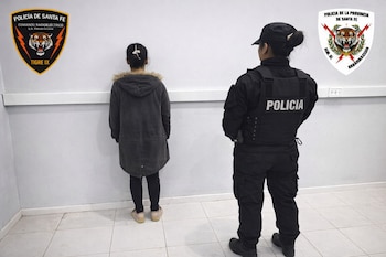 Una persona de espaldas frente a una pared blanca, custodiada por una oficial de policía uniformada de negro y de espaldas. Emblemas policiales en la pared.
