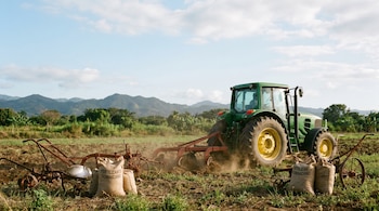 Un tractor verde trabaja la tierra en un campo abierto, con herramientas agrícolas oxidadas y sacos de fertilizante en primer plano. Montañas y vegetación al fondo bajo un cielo claro.