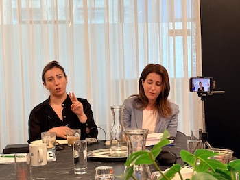 Dos mujeres sentadas en una mesa oscura con vasos y documentos. Una mujer gesticula con la mano; un smartphone la graba. Hay cortinas claras al fondo
