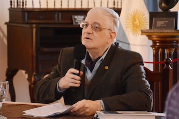 Un hombre con anteojos y saco sostiene un micrófono frente a una mesa con documentos y la bandera de Argentina