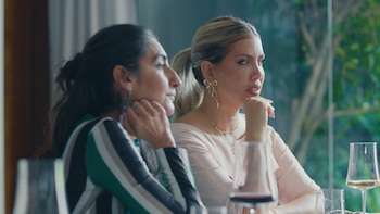 Dos mujeres sentadas a una mesa con copas de vino apoyan la barbilla en sus manos mientras observan hacia un lado
