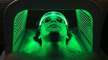 Un hombre recostado con la cara iluminada de verde, usa gafas protectoras y una banda luminosa en la cabeza, debajo de un panel con luces LED verdes.
