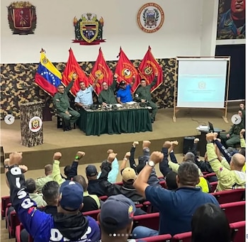 Personas en un auditorio y funcionarios en un estrado con banderas de Venezuela alzan el puño derecho durante una reunión