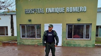 Oficial de Gendarmería Nacional con uniforme y chaleco antibalas frente a la fachada de un hospital verde con el nombre "HOSPITAL ENRIQUE ROMERO"
