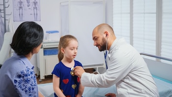 Un médico con bata blanca evalúa el pecho de una niña usando un estetoscopio frente a una mujer en un consultorio