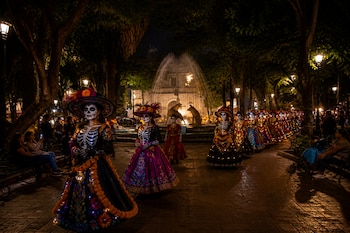 Mujeres caracterizadas de catrinas caminan en fila por una plaza con árboles, farolas y una fuente iluminada durante la noche.